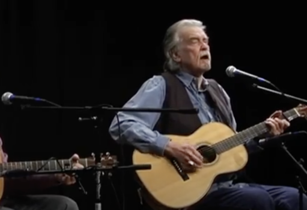 An older man with gray hair and a beard is playing an acoustic guitar and singing into a microphone. He is wearing a blue shirt and a vest. Another person, partially visible in the background, is also playing a guitar. The setting appears to be a stage.