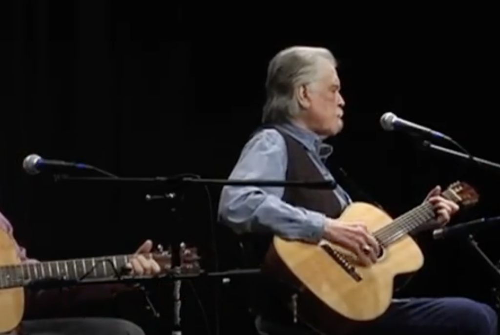 Two musicians seated and playing acoustic guitars, a gray-haired man in the foreground singing into a microphone. The background is dark, focusing attention on the performers and their instruments. Multiple microphones are set up around them.