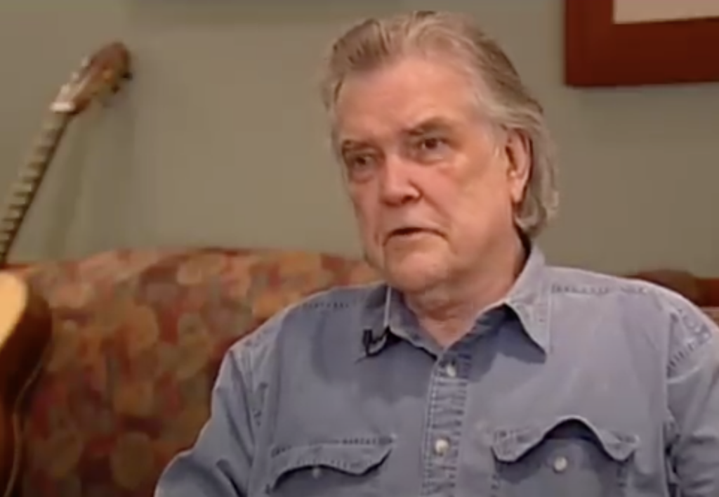 An older man with grey hair and a serious expression sits on a floral patterned couch. He is wearing a denim button-up shirt. A guitar is visible in the background, resting on the couch. The setting appears to be a cozy, indoor living space.