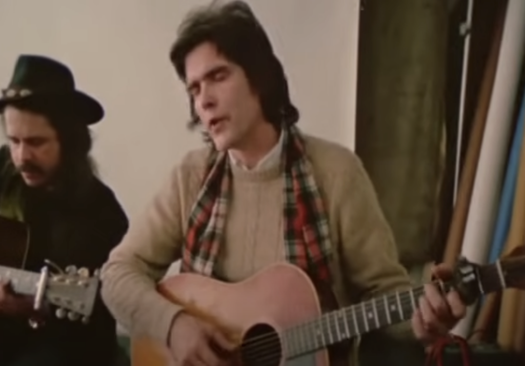 Two musicians sitting side by side, playing acoustic guitars. The musician on the right wears a beige sweater and a checkered scarf, and is singing with closed eyes. The musician on the left wears a black hat and dark clothing, also focused on playing his guitar.