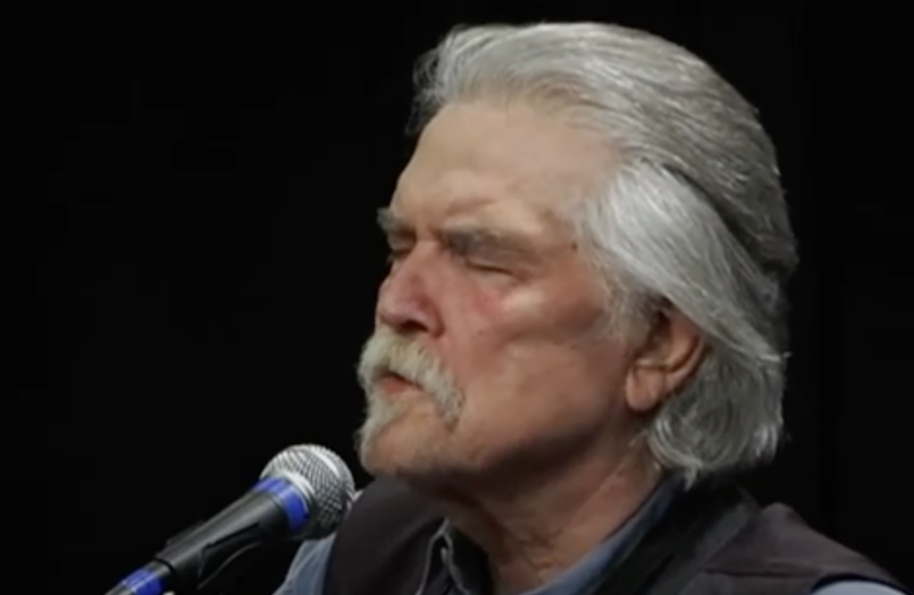 An older man with gray hair and a mustache is singing with his eyes closed into a blue and silver microphone. He is wearing a dark vest over a blue shirt. The background is dark, making him the focal point of the image.