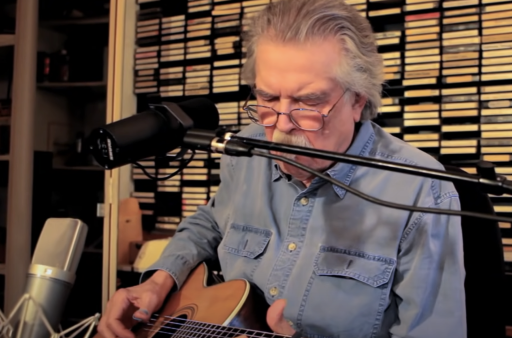 An older man with gray hair and a mustache is playing an acoustic guitar and singing into a microphone. He is seated in a room with shelves filled with numerous audio cassette tapes in the background, wearing a blue denim shirt.