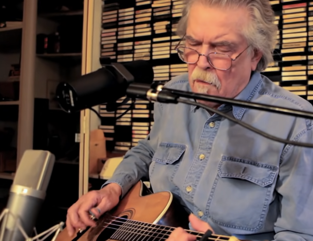 An older man with gray hair and glasses plays an acoustic guitar while singing into a microphone in a cozy, studio-like setting filled with shelves of labeled tapes. He wears a light blue denim shirt.