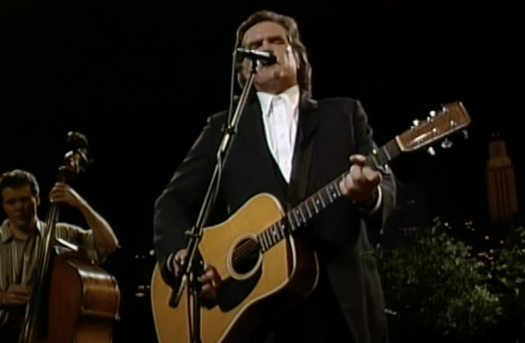 A man in a black suit is singing and playing an acoustic guitar on stage at night. To his left, another musician is playing an upright bass. The background is dark with blurry city lights visible.