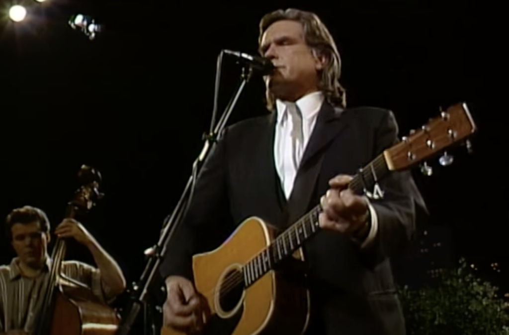 A man in a black suit plays an acoustic guitar and sings into a microphone on stage. Another musician in the background plays a double bass. Bright stage lights shine from above, illuminating the performers. The atmosphere suggests a live music performance.