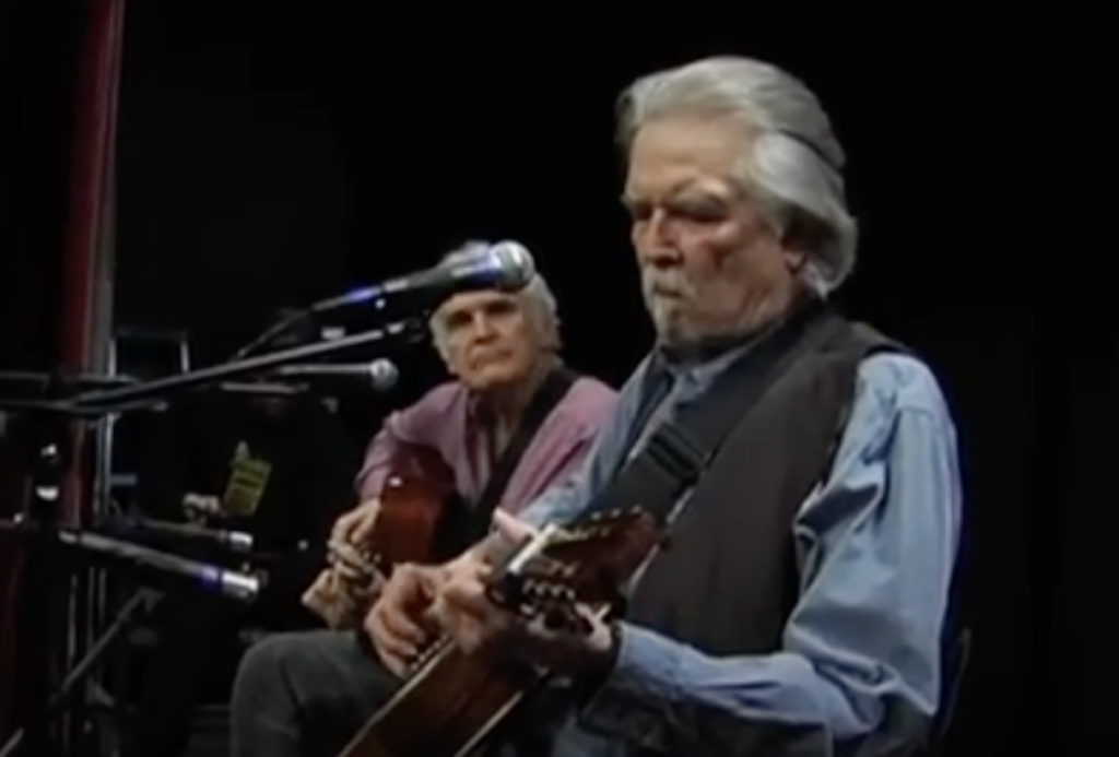 Two elderly men are playing guitars on stage. The man in the foreground has gray hair and is wearing a blue shirt and a black vest, while the man in the background is wearing a pink shirt. Both are focused on their instruments, with a microphone in front of them.