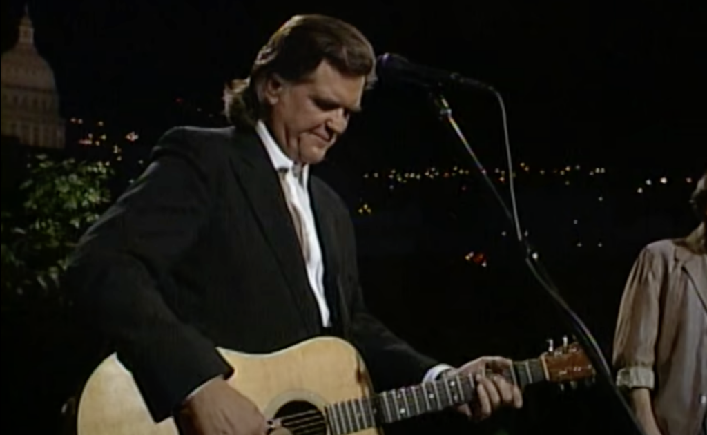 A man in a dark suit is playing an acoustic guitar on stage. He stands in front of a microphone with a background of city lights and a building with a dome. The scene appears to be an evening outdoor performance.