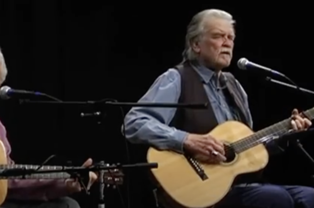 Two elderly men play acoustic guitars while singing into microphones on a dark stage. The man on the right wears a blue shirt, vest, and has white hair and a beard. The man on the left is partially visible in a purple shirt.