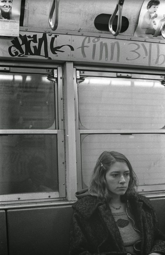 A woman with a somber expression sits alone on a subway train, wearing a coat with a fur collar and a shirt featuring a pair of lips. The background shows graffiti and advertisements above the windows, with reflections slightly visible on the glass. The photo is in black and white.