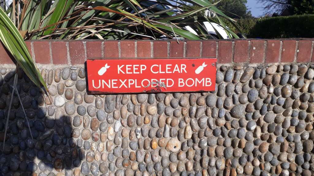 A red warning sign mounted on a pebble-stone wall reads "KEEP CLEAR UNEXPLODED BOMB" in white letters, with two bomb icons on either side of the text. The sign is located beneath some overhanging green plants and a brick ledge.