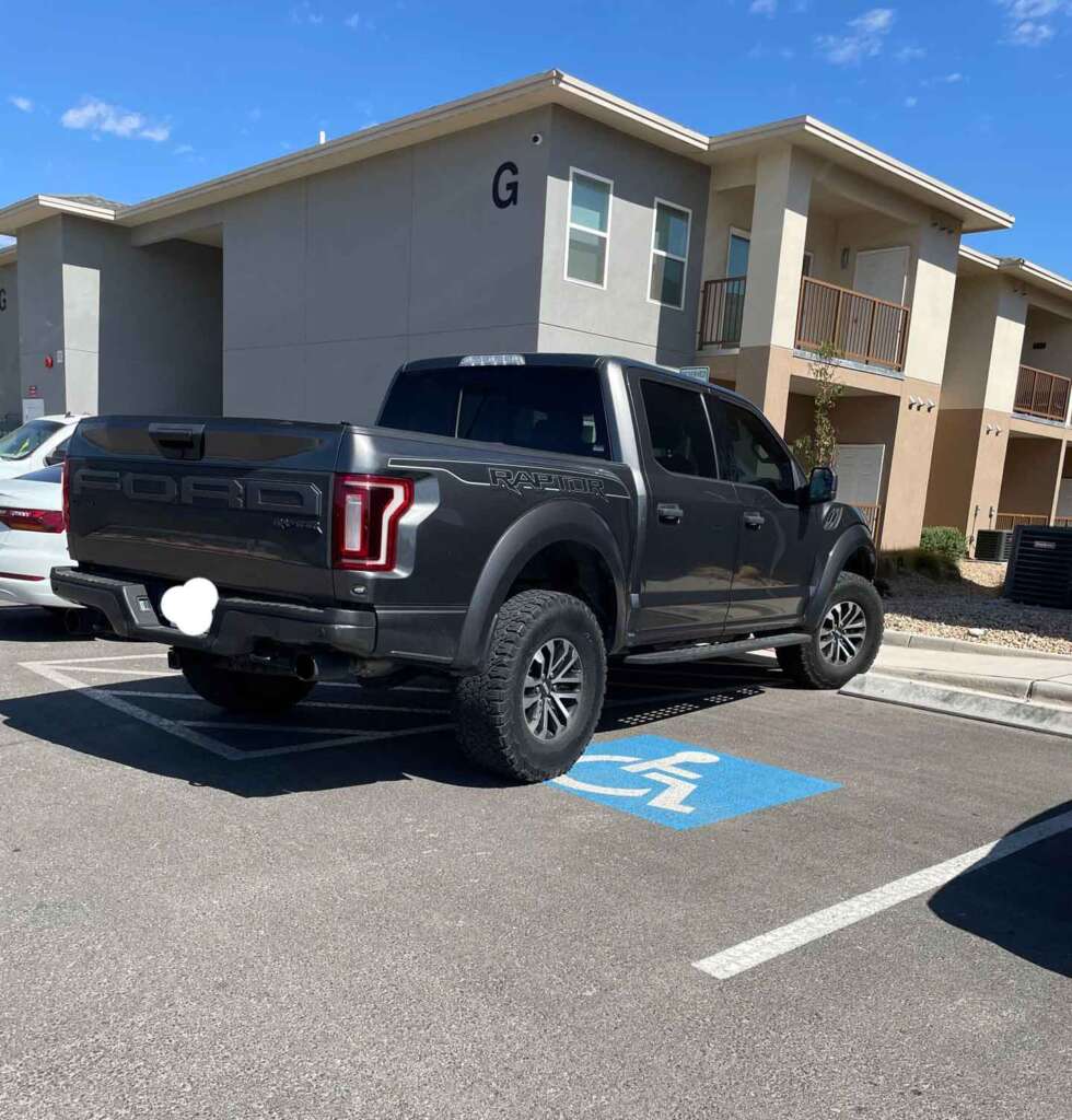 A black Ford Raptor pickup truck is parked in a handicap parking space near an apartment building labeled 'G'. The truck has large wheels and tinted windows. Another car is partially visible parked to the left. The sky is clear and blue.