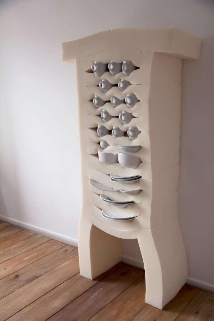 A tall, beige foam furniture piece stands against a white wall on a wooden floor. It has multiple slots holding various white ceramic dishes, cups, and bowls, offering a unique and cushioned storage solution for the items.