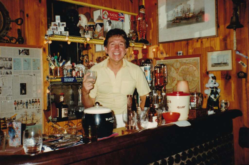 A person stands behind a wooden bar holding a drink and smiling. The bar area is decorated with various items including bottles, a newspaper clipping, a framed ship picture, and novelty items. The background contains wooden walls and dim string lights.