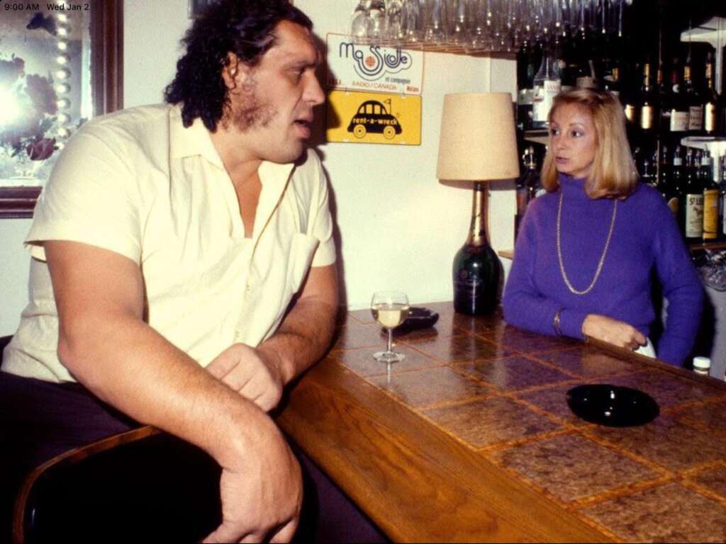 A large man in a white shirt sits at a bar counter talking to a woman in a purple turtleneck sweater. A glass of wine is in front of him. The bar has shelves with glasses and bottles in the background. A poster and a lamp are on the wall behind them.