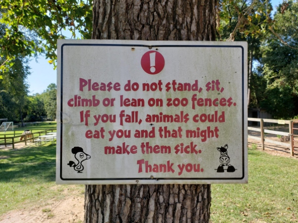 A sign on a tree at a zoo reads: "Please do not stand, sit, climb or lean on zoo fences. If you fall, animals could eat you and that might make them sick. Thank you." The sign includes cartoon images of a goat and a zebra. Trees and a pathway are visible in the background.