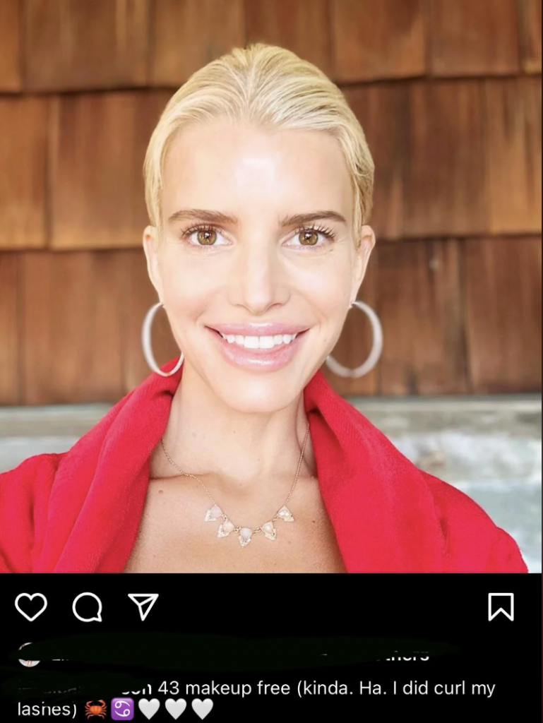 A woman with blonde hair pulled back is smiling at the camera, wearing a red top and a necklace with triangular pendants. She has hoop earrings and minimal makeup. The background features a wooden wall. The social media post caption reads, “43 makeup free (kinda. Ha. I did curl my lashes).”