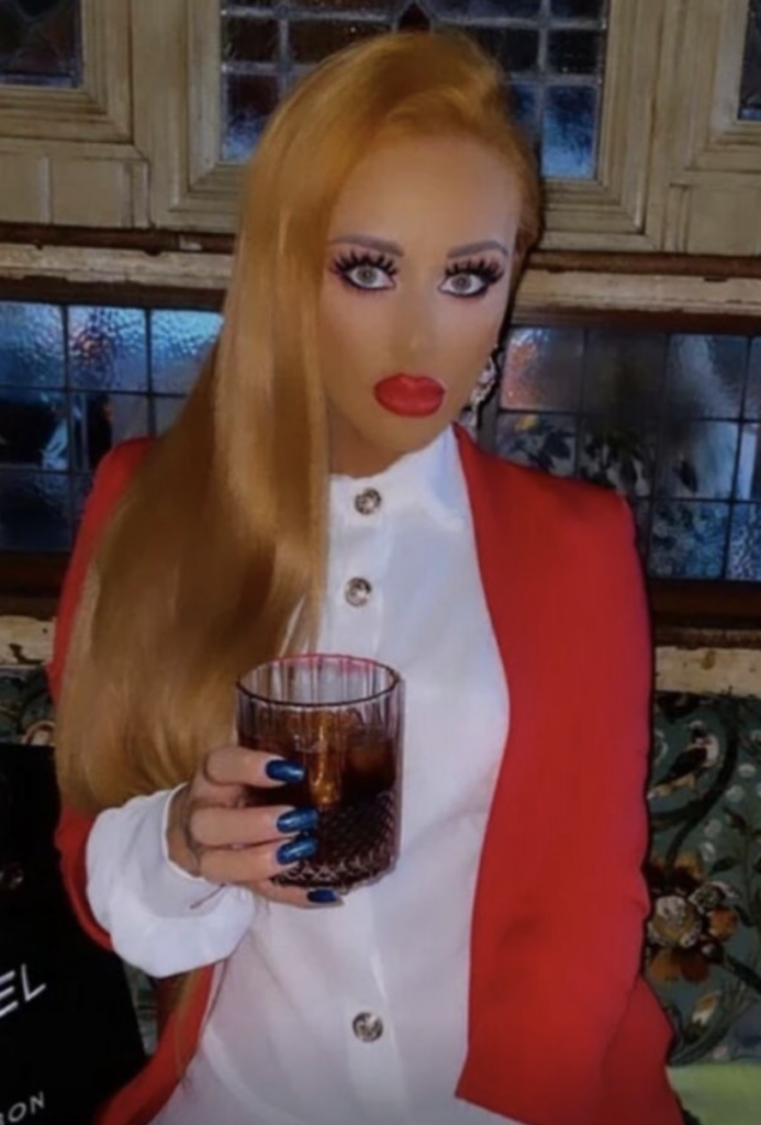 A person with long blonde hair, full makeup, and red lipstick is holding a glass with a dark beverage. They are wearing a red coat over a white buttoned shirt. The background features a tiled section and wooden frames.