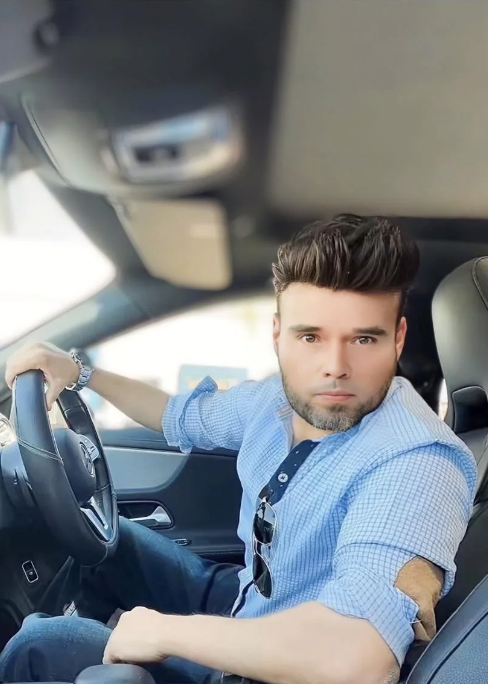 A man with styled dark hair and a short beard sits in the driver's seat of a car, wearing a light blue checkered shirt with rolled sleeves and sunglasses hanging from the collar. He looks directly at the camera with a serious expression and his left hand on the steering wheel.