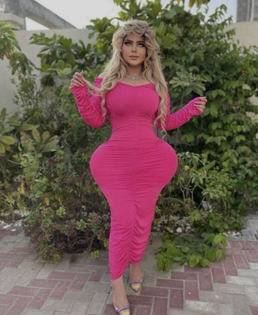 A woman with long blonde hair is wearing a form-fitting, long-sleeved pink dress. She stands outdoors on a paved area with green foliage in the background. She is posed with her arms bent slightly at her sides and wearing open-toed shoes.