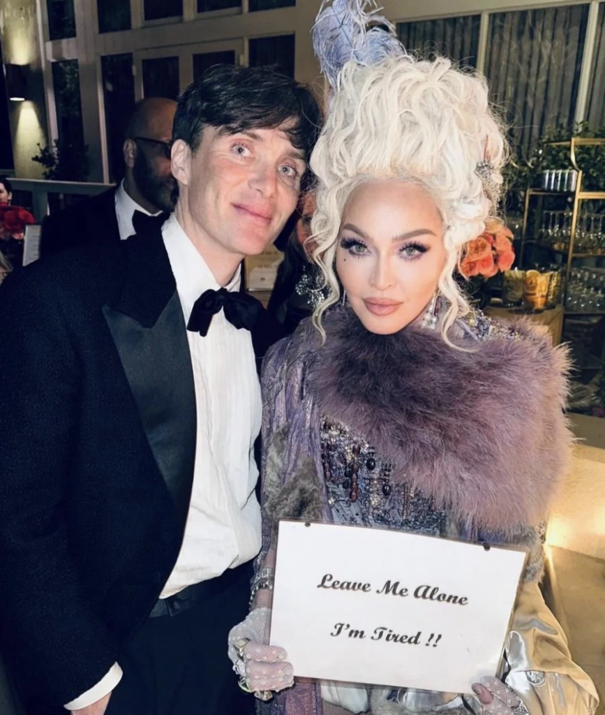 A man in a tuxedo stands next to a woman in elaborate, vintage-style makeup and attire, adorned with feathers and lace. The woman holds a sign reading "Leave Me Alone I'm Tired!!". They are posing indoors at what appears to be a formal event or gathering.