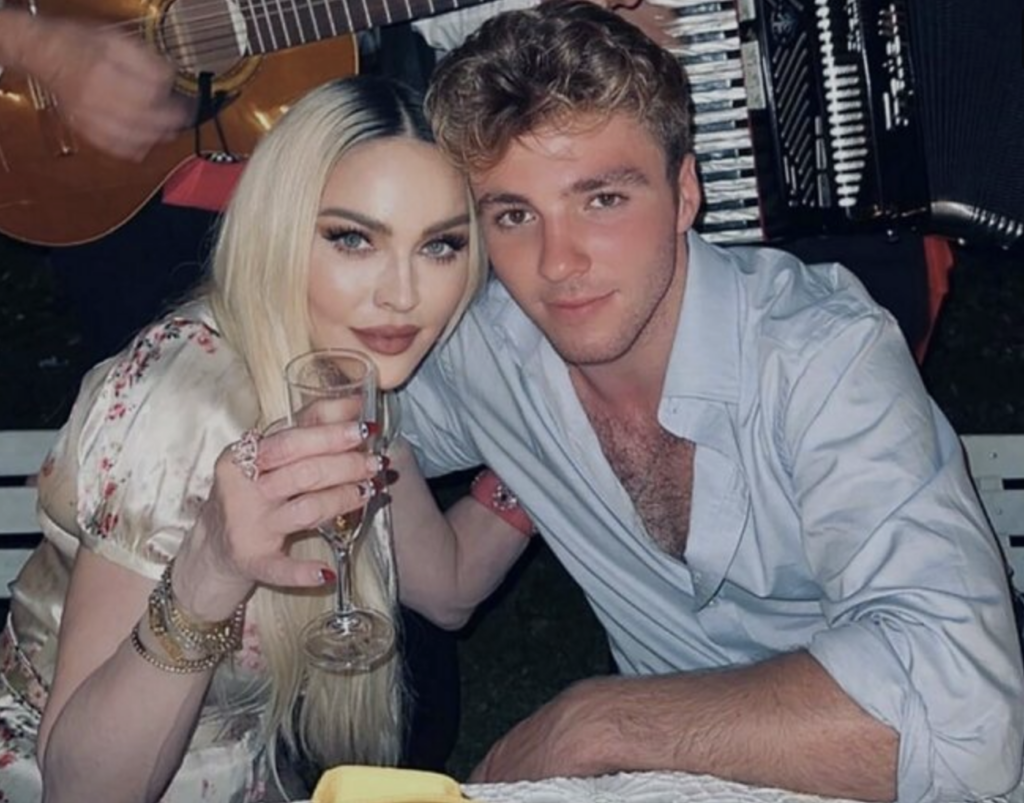 Two people are posing closely together at what appears to be a social event. The person on the left, with long blonde hair, is holding a glass up towards the camera. The person on the right has short brown hair, and they are smiling. Musical instruments are visible in the background.