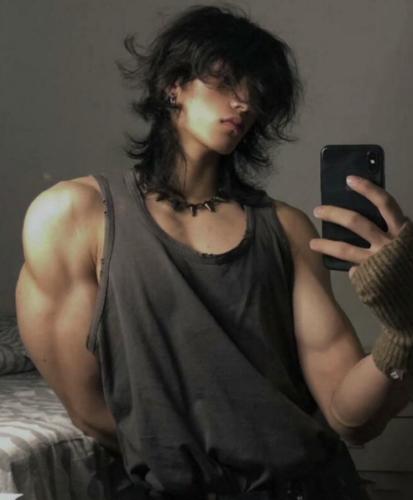 A person with long hair takes a mirror selfie in a dimly lit room. They are wearing a sleeveless dark shirt, a necklace with spikes, and a fingerless glove on their left hand. Their muscular arms are prominently displayed. No facial features are clearly visible.