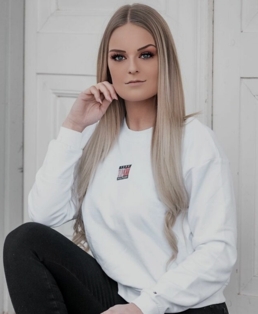 A woman with long, straight blonde hair and makeup sits in front of a white door, resting her elbow on one knee and her hand near her face. She is wearing a white sweatshirt with a small, colorful logo in the center and black pants.