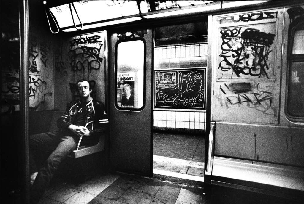 Black and white photo of a man sitting alone on a bench inside a graffiti-covered subway car. Another train is visible outside through the open doors, and the tiled subway platform walls show more graffiti. The atmosphere feels gritty and urban.