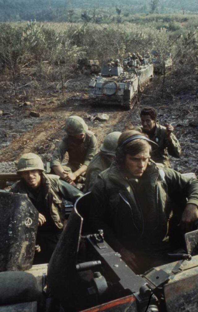 Soldiers in military uniforms and helmets ride atop armored vehicles in a rugged, wooded area. The leader in the foreground wears communication gear, while others around him hold weapons and various equipment, indicating a wartime or training scenario.