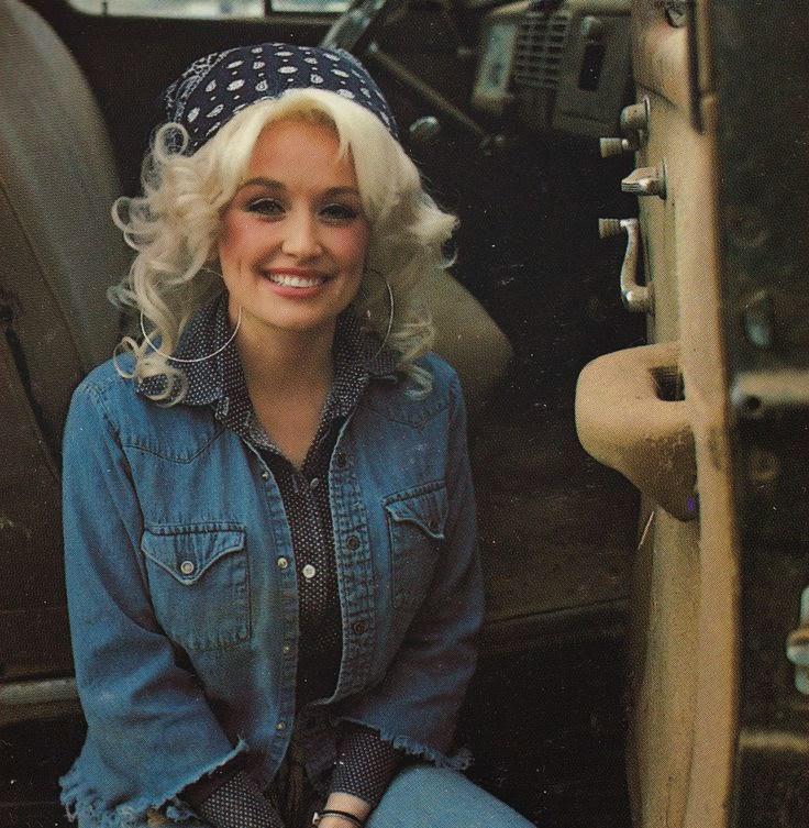 A woman with curly blonde hair, wearing a blue bandana, denim jacket, and hoop earrings, smiling while sitting inside a vehicle. The interior of the vehicle appears rustic, with visible controls and textures.