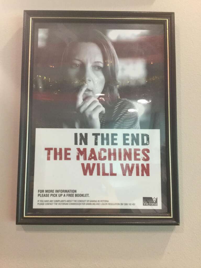 A framed poster shows a concerned woman with her hand on her chin. The text on the poster reads, "IN THE END THE MACHINES WILL WIN." Below, there's smaller text with information on where to get more details and help regarding gaming in Victoria, Australia.