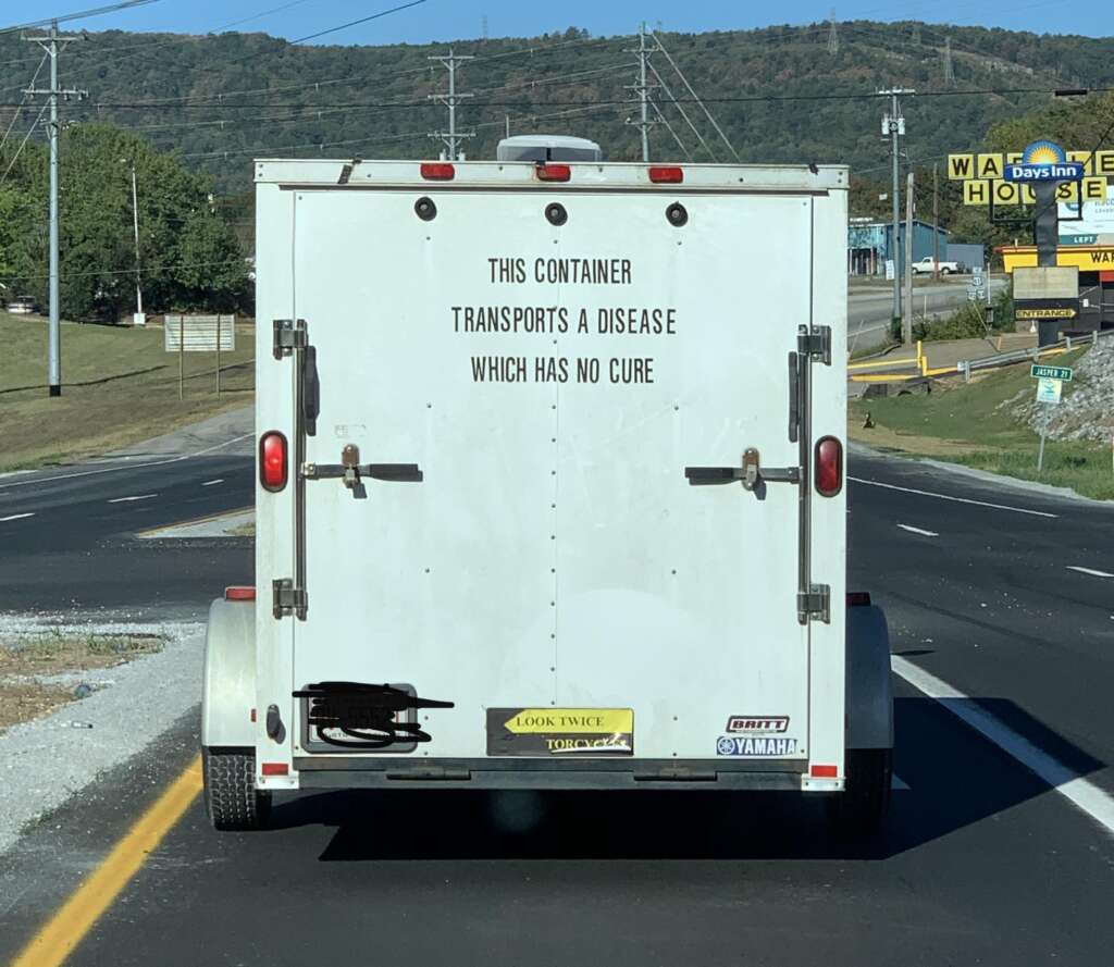 A white enclosed trailer on a road has the text "THIS CONTAINER TRANSPORTS A DISEASE WHICH HAS NO CURE" on its rear door. The trailer is secured with a padlock and features various stickers, including "LOOK TWICE," "TOY HAULER," and "YAMAHA.