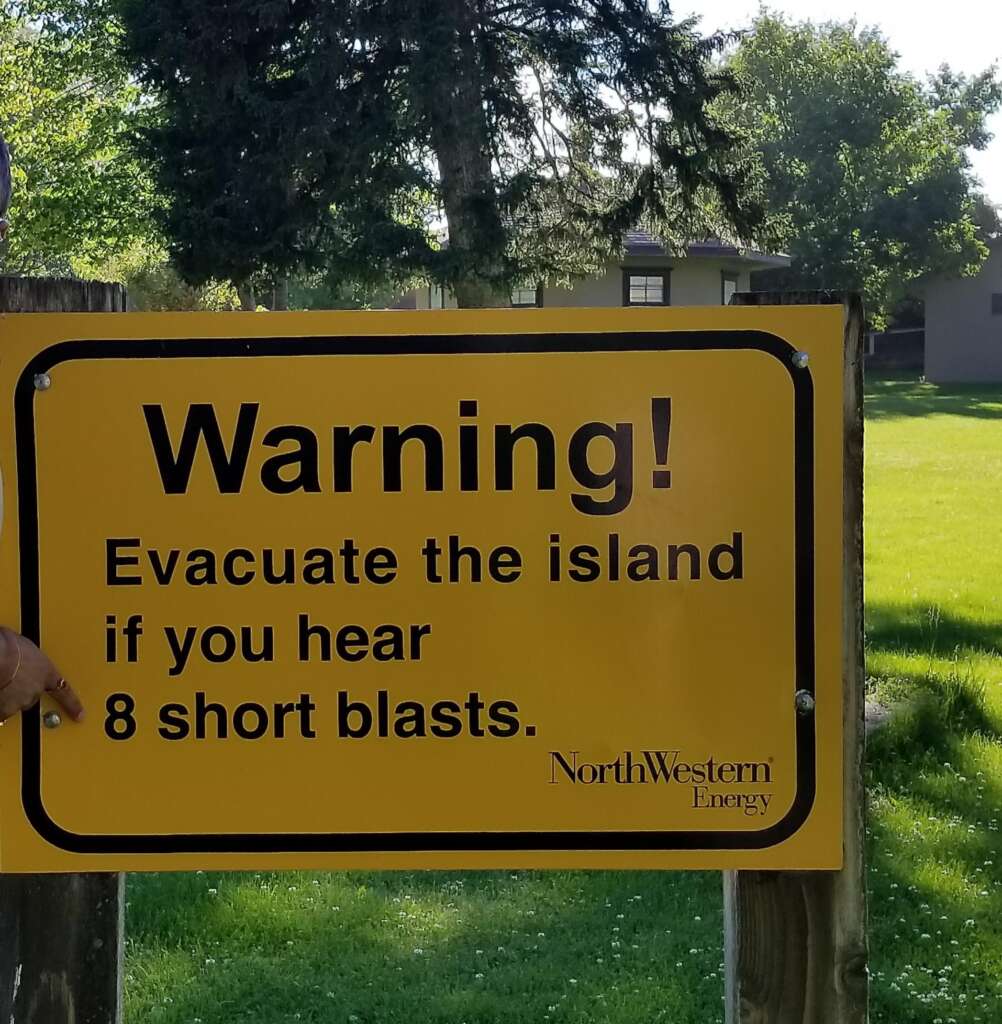 A warning sign posted on a wooden fence reads, "Warning! Evacuate the island if you hear 8 short blasts," with the NorthWestern Energy logo in the bottom-right corner. The background is a grassy area with trees and a house visible.