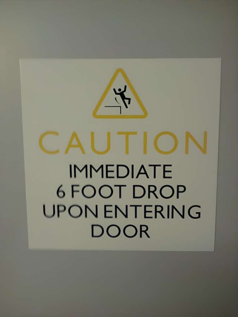 A sign with a yellow triangle warning symbol and text that reads, "CAUTION IMMEDIATE 6 FOOT DROP UPON ENTERING DOOR." The illustration within the triangle shows a person falling. The sign is displayed on a plain surface.