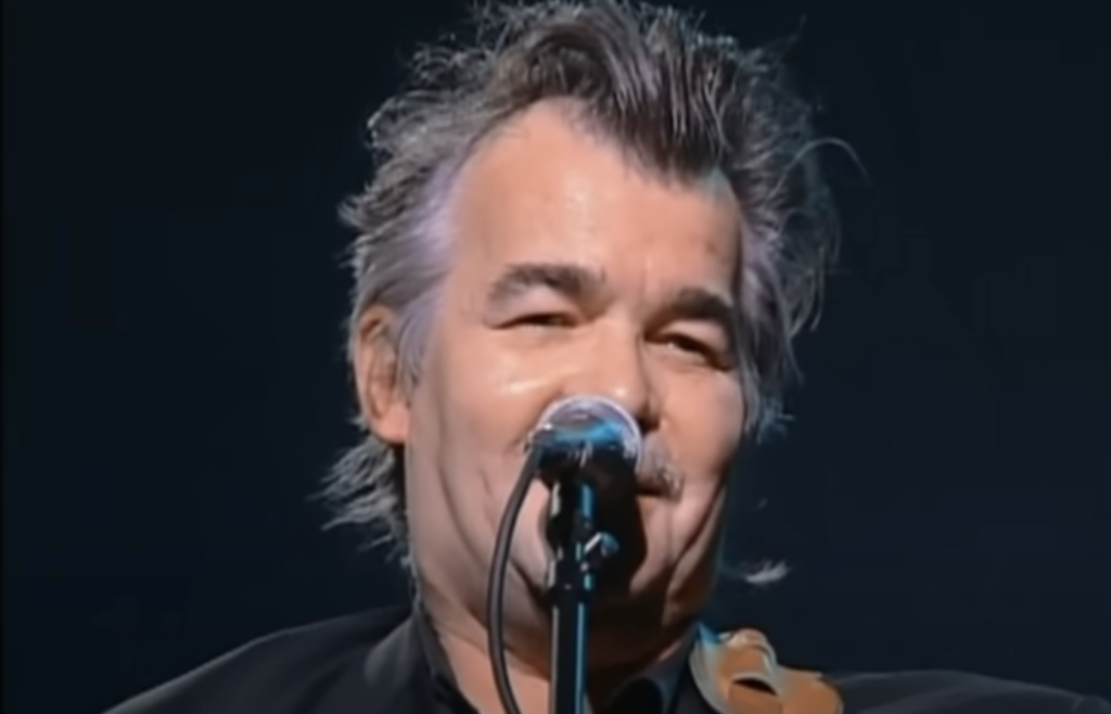 A middle-aged person with grey hair performs on stage, holding a microphone close to their mouth. They are dressed in a dark-colored outfit, and the background is dark, emphasizing the person’s face and expression.