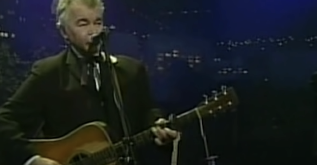 A person with gray hair is playing an acoustic guitar and singing into a microphone on a dimly lit stage. The background features blurry city lights. The person is wearing a dark suit and tie.