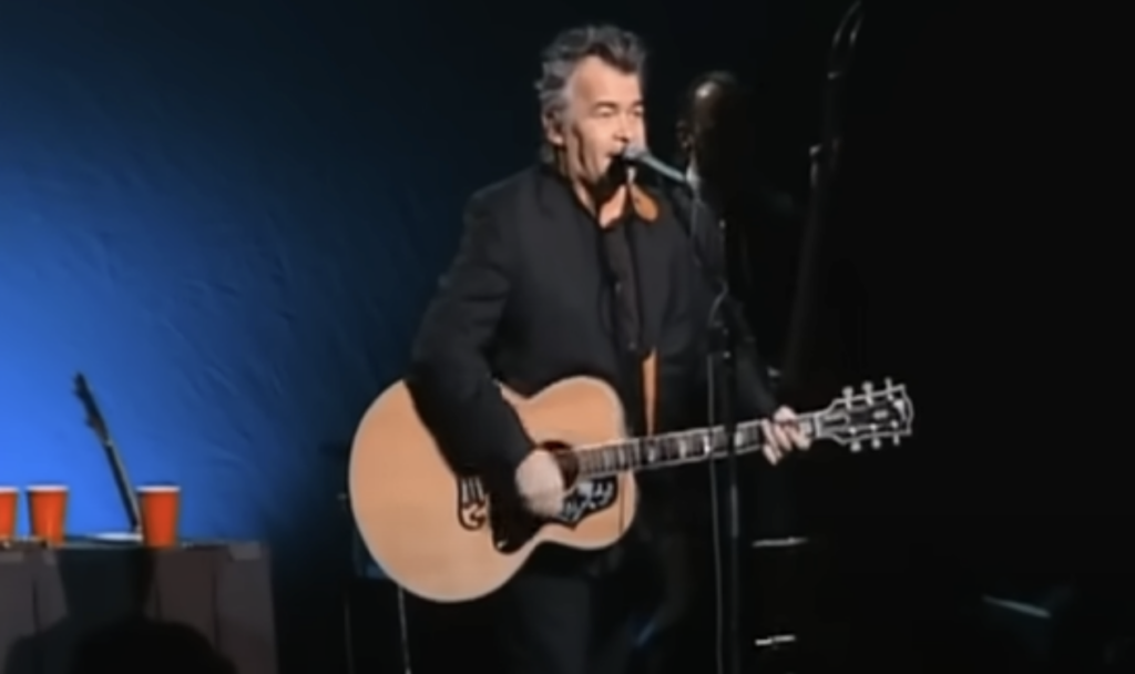 A man with gray hair is playing an acoustic guitar and singing into a microphone on a stage. He is dressed in a black suit. There is a dark blue backdrop behind him and a table with red cups to his left. The stage lighting is low, focusing on the performer.