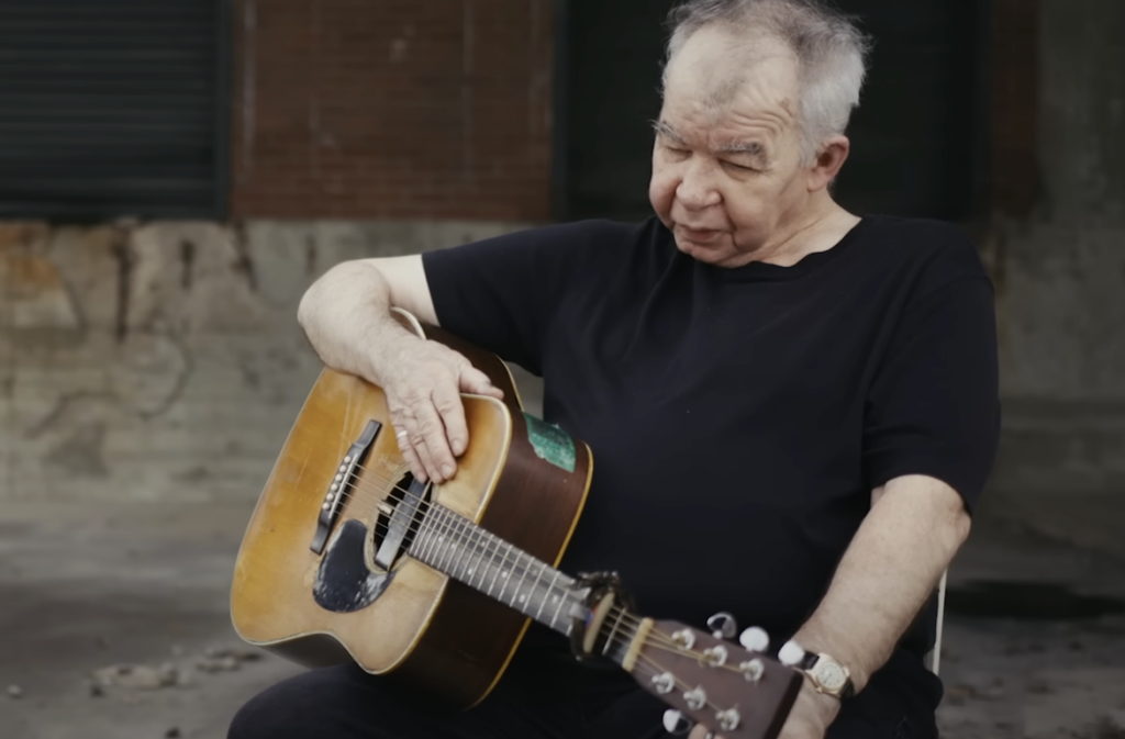 An elderly man with gray hair sits holding an acoustic guitar. He is casually dressed in a black t-shirt and is outdoors against a backdrop of an old, weathered building. His expression appears thoughtful as he looks down at his guitar.