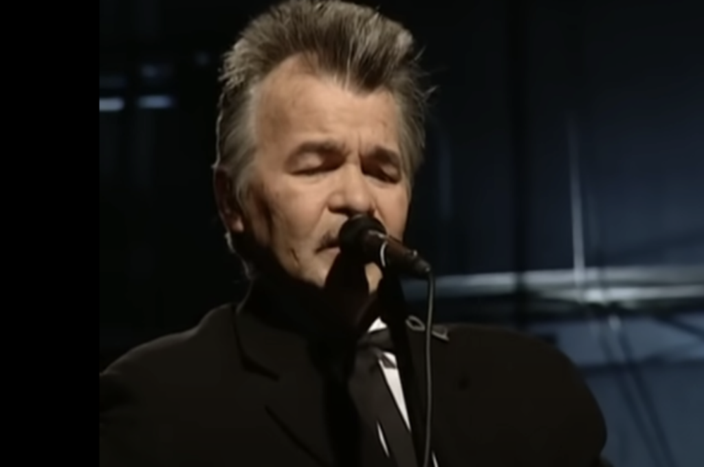 An older man with gray hair and sideburns is singing into a microphone. He is wearing a black suit and a tie. The background is dimly lit, with soft focus suggesting an indoor stage setting.