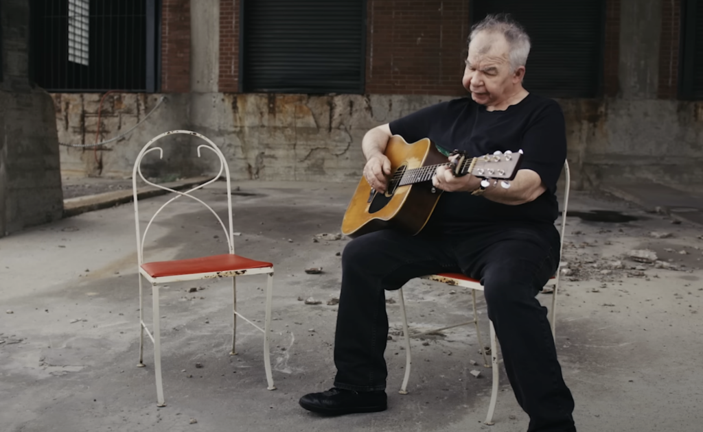 An elderly man with white hair is sitting on a chair in a desolate, industrial setting, playing an acoustic guitar. He is dressed in black, and there is an empty chair with a red seat next to him. The ground is worn, with debris scattered around.
