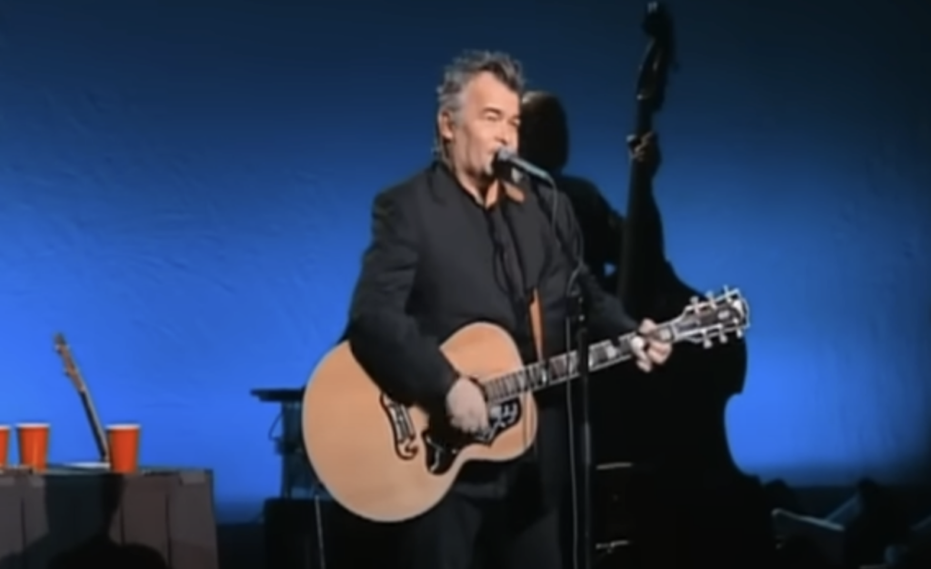 A man with gray hair, dressed in a black suit, is performing on stage while playing an acoustic guitar and singing into a microphone. The stage background is blue, and there is a double bass and other instruments visible in the dimly lit background.