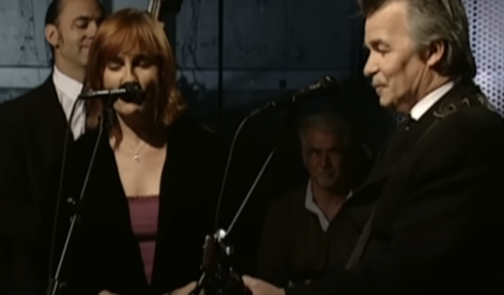 A man and a woman are performing on stage, both singing into microphones. The man is playing a guitar. There are additional musicians in the background, including a man with a double bass. The woman has red hair and is wearing a dark outfit. The background is dimly lit.
