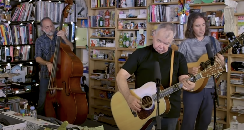 A trio of musicians performs in a cozy, cluttered room filled with shelves of books and various items. The front musician plays an acoustic guitar and sings, while the others play a double bass and another guitar. Microphones are set up in front of them.