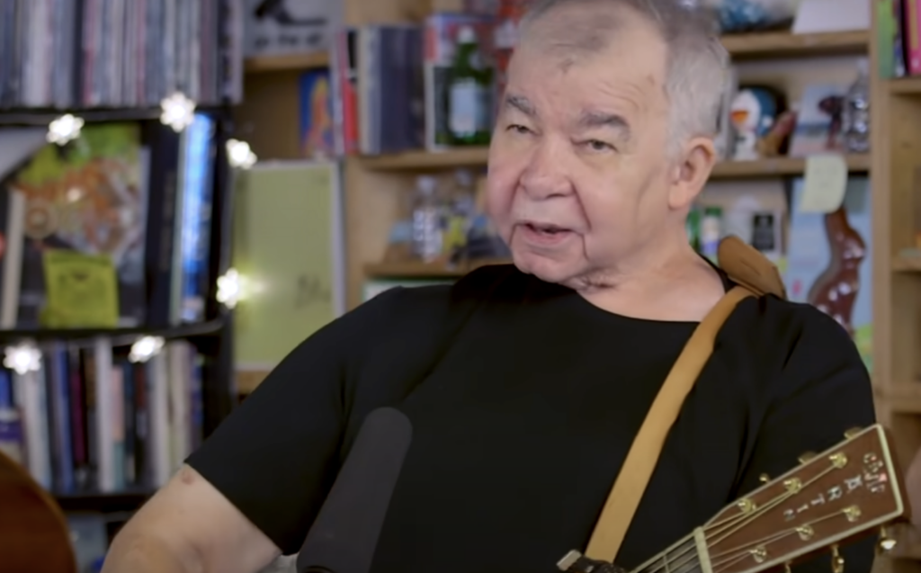 An older man with gray hair is playing an acoustic guitar. He is wearing a black t-shirt and a brown strap for the guitar. Behind him, shelves are filled with books and various items, and some string lights add a decorative touch.