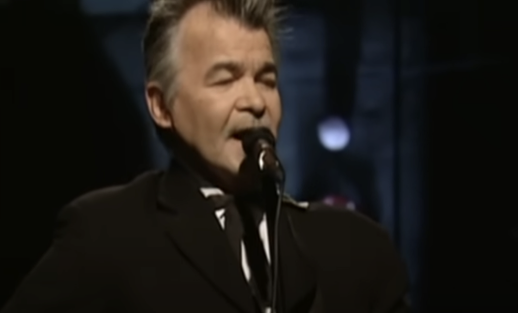 A person with gray hair and a mustache is singing into a microphone. The individual is wearing a dark suit and is performing on stage with a dark background and faint, out-of-focus lights behind them.