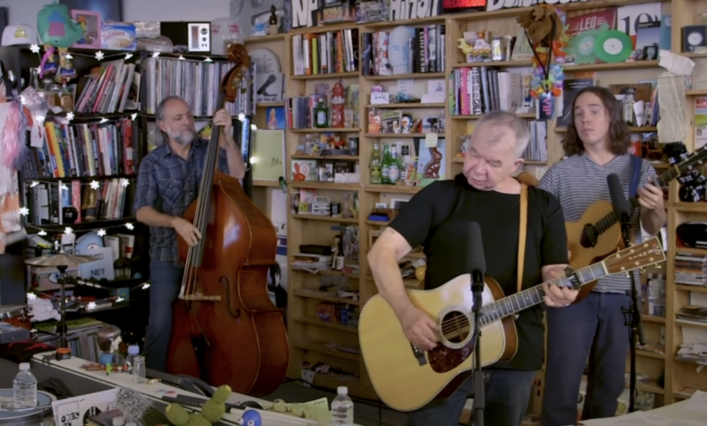 A small band performs in a cozy, cluttered room. A man with a double bass, another sings and plays an acoustic guitar, and the third musician watches while playing a guitar. The background is filled with books, musical equipment, and various knick-knacks.