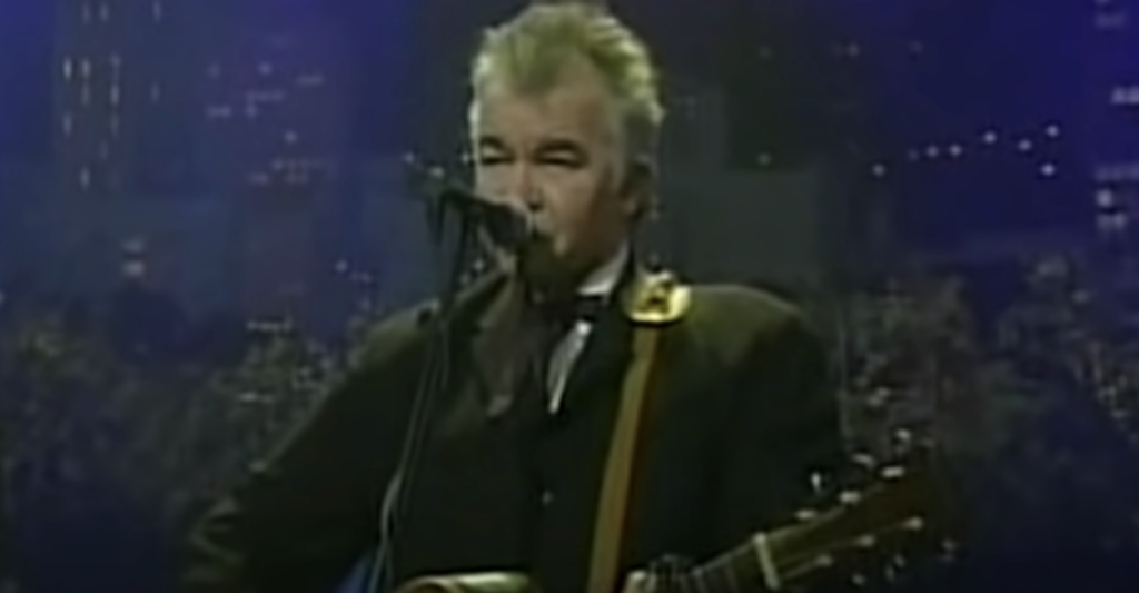 A man with gray hair is singing into a microphone while playing an acoustic guitar. He is wearing a dark-colored suit. Behind him, the background is dark and blurred, possibly showing a nighttime cityscape.