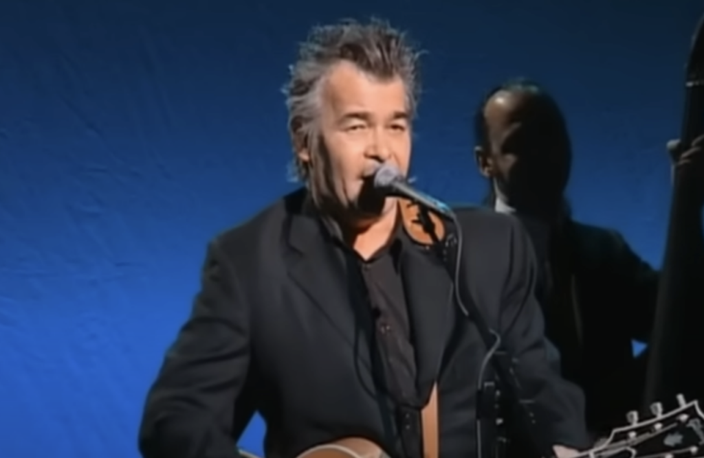 A man with gray hair and a black suit sings into a microphone while strumming an acoustic guitar. He stands in front of a blue backdrop. A second person, holding an upright bass, is in the background, partially obscured.