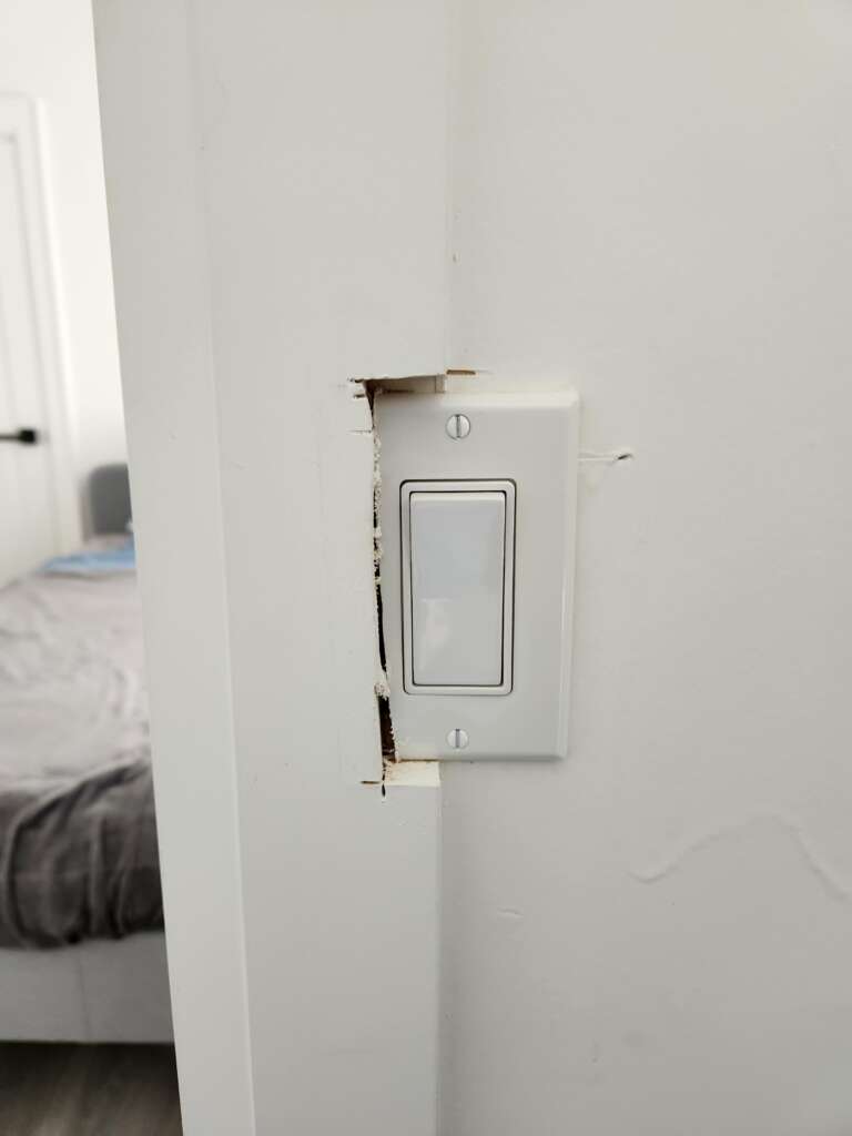 A white light switch is installed on a damaged wall with some visible cracks and chipped paint around it. In the background, there is a bed with gray bedding in a room. The door to the room is partially visible on the left side.