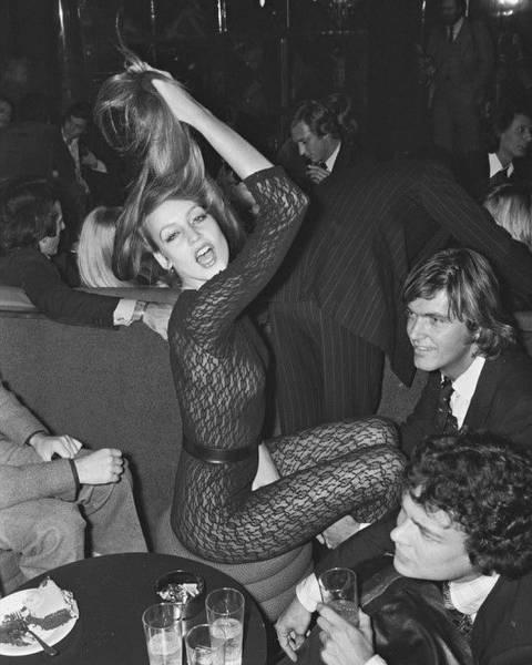 A woman in a sheer, patterned bodysuit energetically poses while holding her hair above her head in a lively nightclub setting. She is surrounded by seated individuals, some holding drinks, and the atmosphere appears vibrant and celebratory.